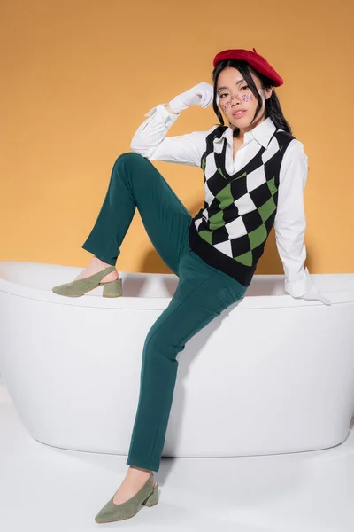 Stylish asian model in beret and gloves posing on bathtub on orange background — Stock Photo