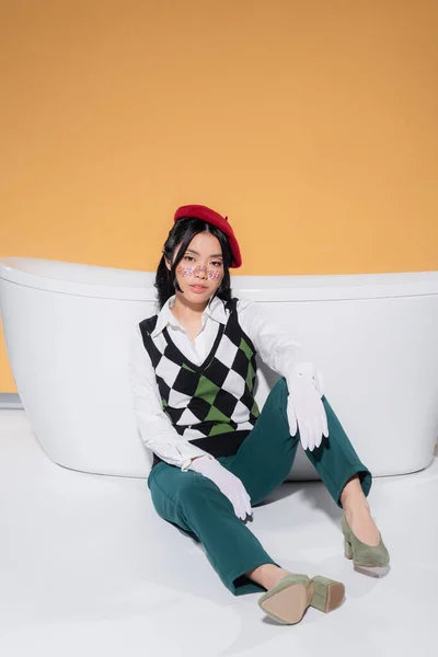 Fashionable asian woman in beret and gloves sitting near bathtub on orange background — Stock Photo