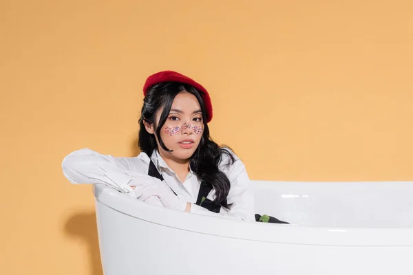 Elegante mujer asiática en guantes y boina sentada en bañera sobre fondo naranja - foto de stock