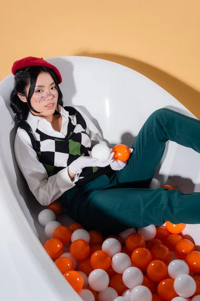 Vue grand angle de la femme asiatique élégante tenant des boules tout en étant assis dans la baignoire sur fond orange — Photo de stock