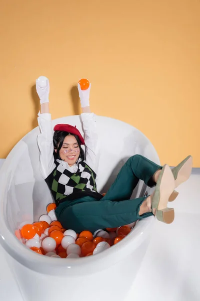 Positive asian model in vintage clothes holding balls in bathtub on orange background — Stock Photo