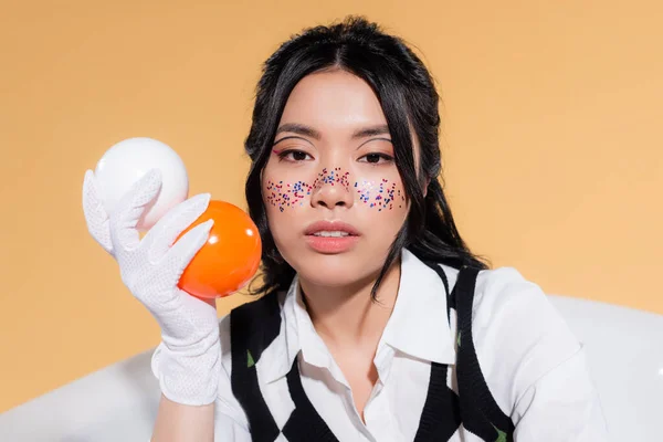 Ritratto di giovane modella asiatica con brillantini sul viso tenendo le palle e guardando la fotocamera nella vasca isolata su arancione — Foto stock
