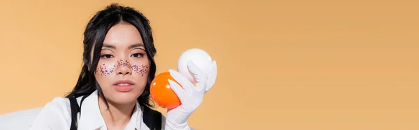 Stylish asian woman with glitter makeup holding balls isolated on orange, banner — Stock Photo