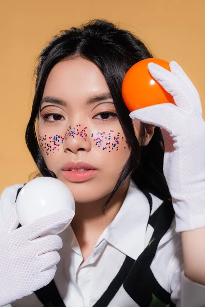 Portrait de modèle asiatique avec visage pailleté tenant des boules et regardant la caméra isolée sur orange — Photo de stock