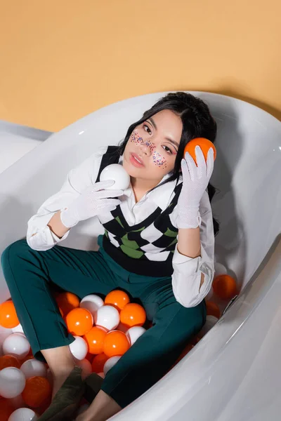 Vue grand angle du modèle asiatique tendance dans des gants tenant des boules tout en étant assis dans la baignoire sur fond orange — Photo de stock