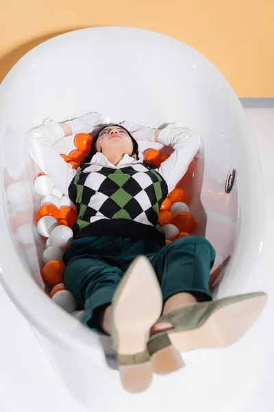 Elegante modello asiatico in guanti sdraiato nella vasca da bagno con palle su sfondo arancione — Foto stock