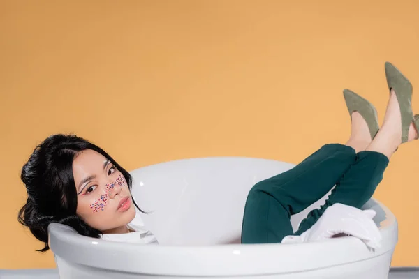 Modelo asiático bonito com brilho no rosto olhando para a câmera enquanto sentado na banheira no fundo laranja — Fotografia de Stock