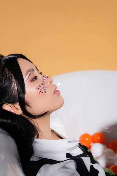 Side view of trendy asian woman with glitter makeup sitting in blurred bathtub with balls isolated on orange — Stock Photo