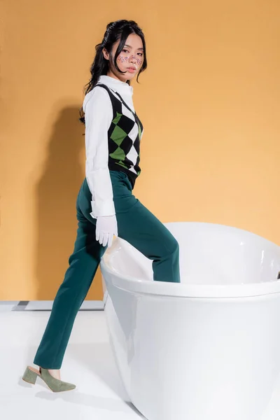 Trendy modello asiatico passo nella vasca da bagno e guardando la fotocamera su sfondo arancione — Foto stock