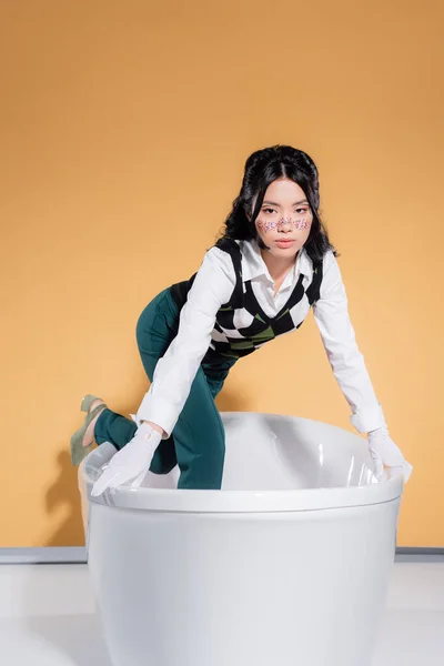 Stilvolle asiatische Frau schaut in die Kamera, während sie in der Badewanne auf orangefarbenem Hintergrund steht — Stockfoto