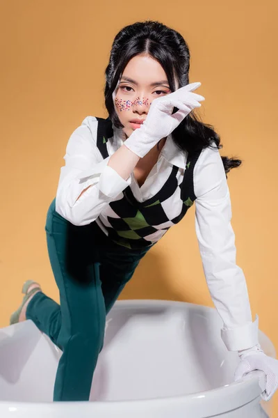 Stylish asian model in gloves looking at camera in bathtub on orange background — Stock Photo