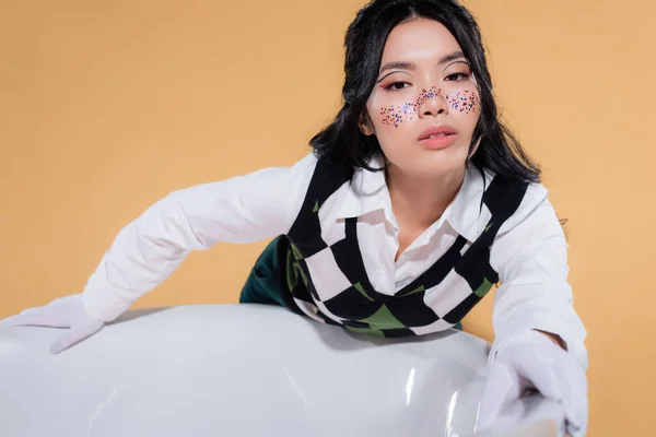 Trendy young asian model with glitter makeup looking at camera near bathtub isolated on orange — Stock Photo