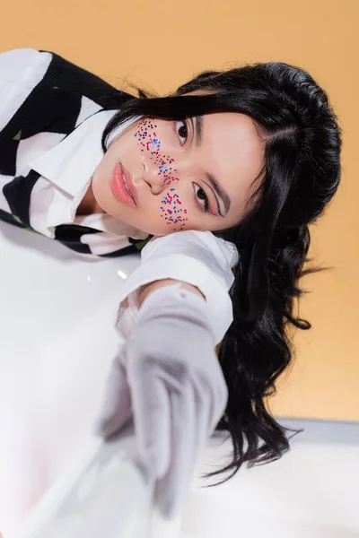 Retrato de modelo bastante asiático con maquillaje de brillo tumbado en la bañera sobre fondo naranja - foto de stock