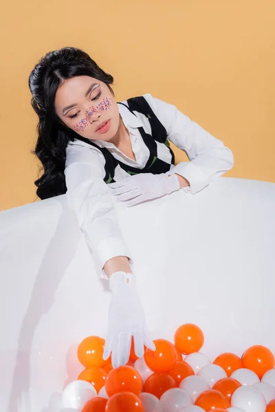 Modelo asiático de moda en guantes mirando bolas en bañera aislado en naranja - foto de stock