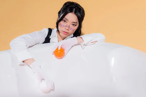Ritratto di modello asiatico alla moda in guanti che tengono le palle vicino alla vasca da bagno su sfondo arancione — Foto stock