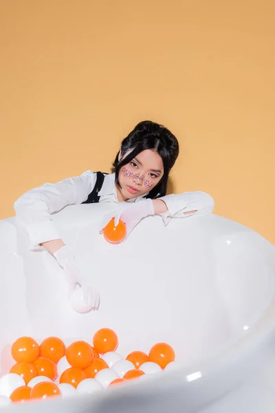 Jolie femme asiatique avec des paillettes sur le visage tenant des boules près de la baignoire sur fond orange — Photo de stock