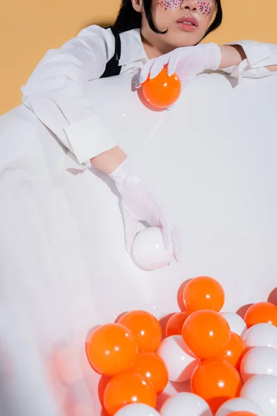 Cropped view of young stylish woman in gloves holding balls near bathtub on orange background — Stock Photo