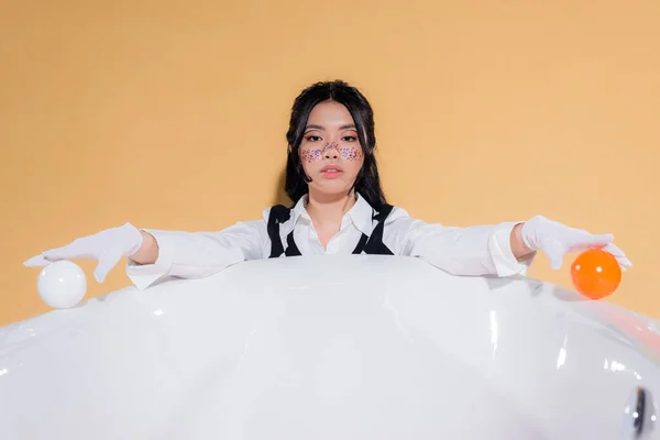 Modelo asiático de moda en guantes sosteniendo bolas cerca de la bañera aislado en naranja - foto de stock