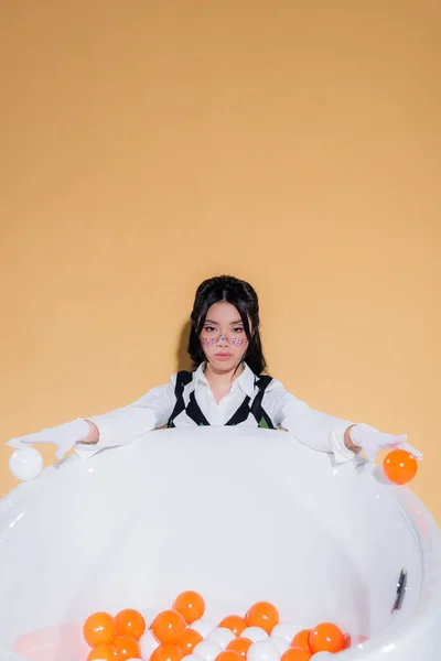 Joven modelo asiático con brillo maquillaje celebración bolas cerca de bañera aislado en naranja - foto de stock