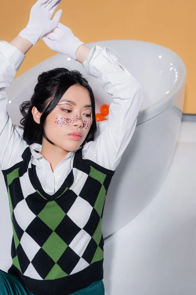 Trendy asian model in gloves sitting near bathtub with balls on orange background — Stock Photo