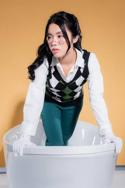 Donna asiatica alla moda in guanti in piedi nella vasca da bagno su sfondo arancione — Foto stock