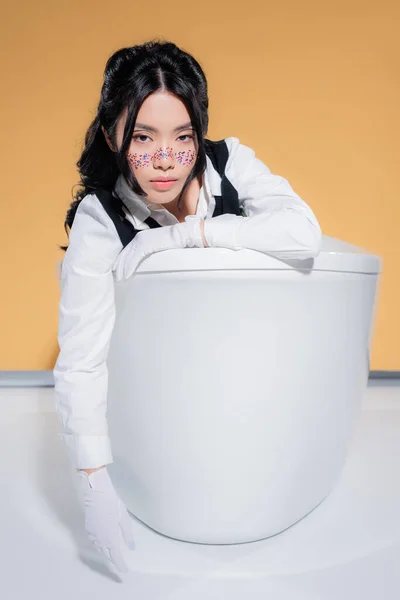 Elegante mujer asiática en guantes mirando a la cámara en bañera sobre fondo naranja - foto de stock