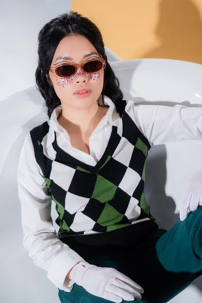 Asian model in sunglasses and retro clothes sitting in bathtub on orange background — Stock Photo