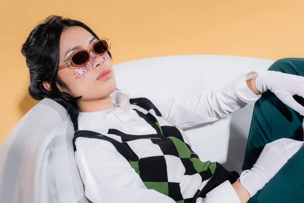 Trendy modello asiatico in occhiali da sole e guanti seduto nella vasca da bagno su sfondo arancione — Foto stock