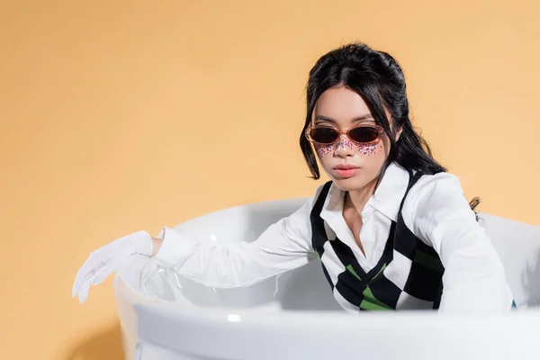 Femme asiatique à la mode avec des lunettes de soleil et des paillettes sur le visage en regardant la caméra dans la baignoire sur fond orange — Photo de stock