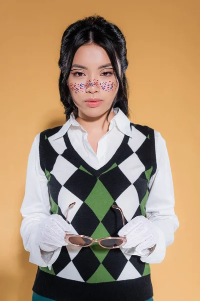 Portrait of stylish asian woman in gloves holding sunglasses on orange background — Stock Photo