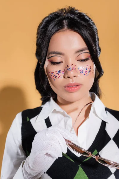 Jeune modèle asiatique avec des paillettes sur le visage tenant des lunettes de soleil sur fond orange — Photo de stock