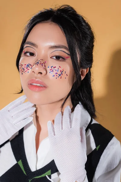 Modisches asiatisches Modell mit Glitzern im Gesicht, das in die Kamera auf orangefarbenem Hintergrund schaut — Stock Photo