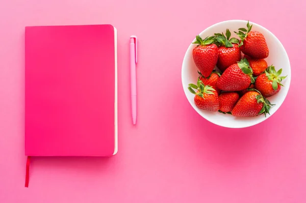 Notebook perto de caneta e morangos inteiros em tigela no fundo rosa — Fotografia de Stock