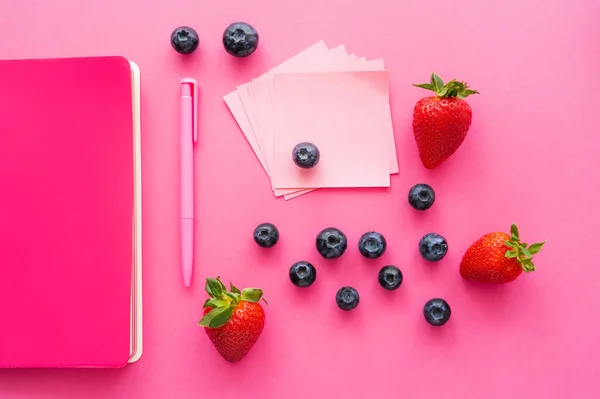 Vista dall'alto di bacche vicino a note appiccicose e notebook sulla superficie rosa — Foto stock
