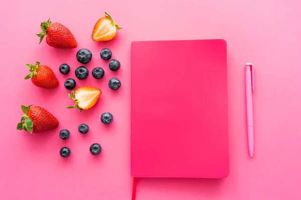 Top view of natural berries near notebook and pen on pink background — Stock Photo