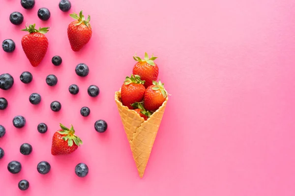 Poser à plat avec des fraises dans un cône de gaufre et des bleuets sur fond rose — Photo de stock