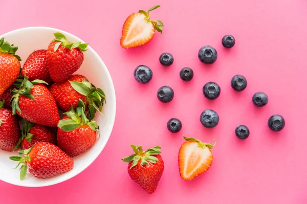 Vue du dessus des fraises biologiques dans un bol près des bleuets sur fond rose — Photo de stock
