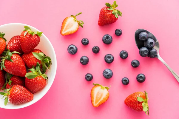 Acostado plano con fresas y arándanos cerca de cuenco y cuchara sobre fondo rosa - foto de stock