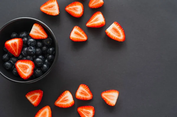 Top view of cut strawberries near blueberries in bowl on black background — Stock Photo