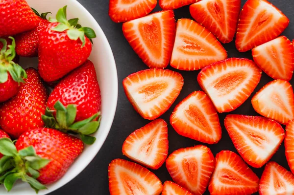 Flache Lage mit geschnittenen und ganzen Erdbeeren in Schale auf schwarzem Hintergrund — Stockfoto