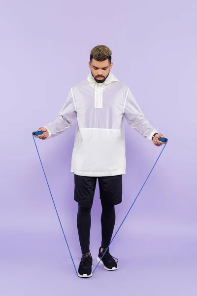 Full length of young african american man in trendy sportswear exercising with skipping rope on purple — Stock Photo