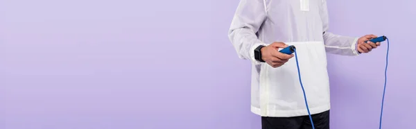 Cropped view of african american man in trendy sportswear holding skipping rope isolated on purple, banner — Stock Photo