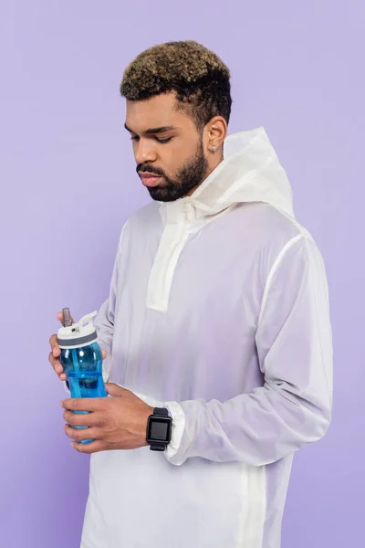 Joven afroamericano hombre en ropa deportiva celebración botella de deportes aislado en púrpura - foto de stock