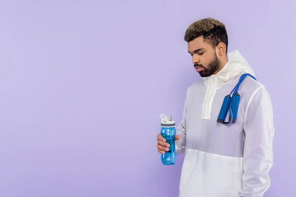 Afro-americano em sportswear com pular corda segurando esportes garrafa isolada em roxo — Fotografia de Stock