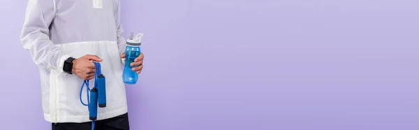 Vue recadrée d'un sportif afro-américain tenant une bouteille de sport et une corde sautante isolée sur du violet, bannière — Photo de stock