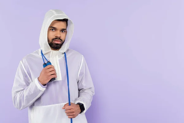 African american sportsman in stylish sportswear with hood looking at camera and holding skipping rope isolated on purple — Stock Photo