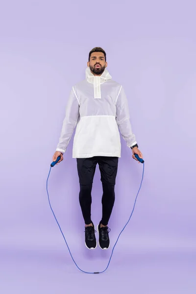 Full length of african american sportsman in sportswear jumping with skipping rope on purple — Stock Photo