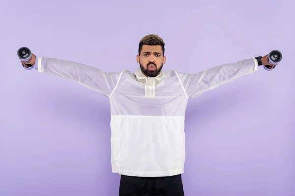Бородатий афроамериканський спортсмен з простягнутими руками вправами з гантелями на фіолетовому — стокове фото