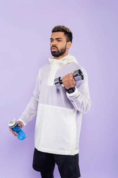 Bearded african american sportsman exercising with dumbbell and holding sports bottle isolated on purple — Stock Photo