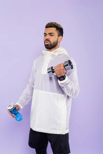 Sportif afro-américain barbu faisant de l'exercice avec haltère et tenant bouteille de sport isolé sur violet — Photo de stock